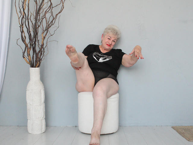 KayleSmoll, elderly woman with short white hair, black shirt, and black panties, sits on a white chair, raising one leg, showing her bare foot and pink panties.