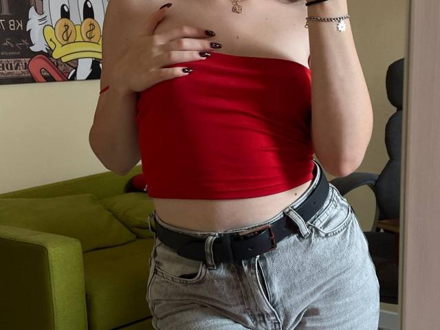 Valissia, vêtue d'un haut rouge sans manches qui couvre ses seins avec une main, porte un jean gris avec une ceinture noire et du vernis à ongles noir, dans un cadre intérieur décontracté..