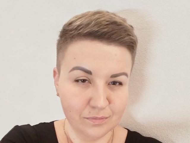 RachelExcite has short brown hair, thick eyebrows, and a slight smile. She's wearing a black top with a gold necklace, looking directly at the camera.