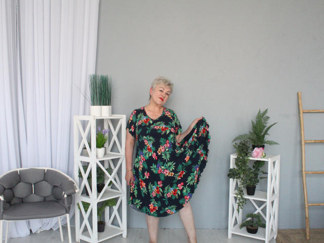 KayleSmoll, short blonde hair, red lipstick, black floral dress, holding skirt, standing between white shelves with plants, gray chair on left, wooden ladder on right.