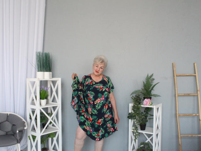 KayleSmoll with short gray hair, wearing a floral dress, holding it up, showing her ass, standing between white shelves with plants and a gold ladder.