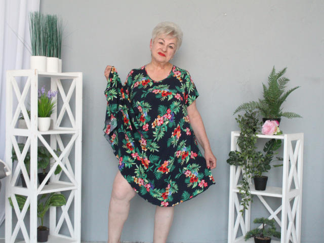 KayleSmoll with short gray hair, red lipstick, wearing a black floral dress, holding up the skirt, standing between two white shelves with plants. Her ass is slightly visible.