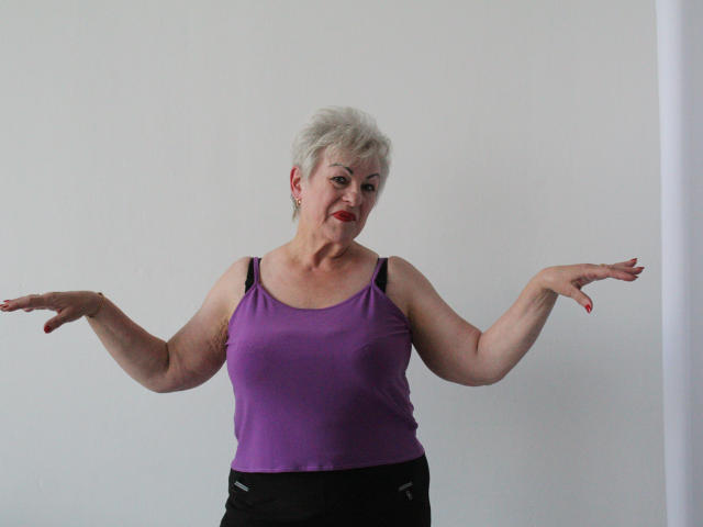 KayleSmoll, middle-aged woman with short white hair, purple tank top, black bra, and black pants, arms spread, red lipstick, slight smile, standing against plain white wall.
