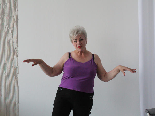 KayleSmoll has short white hair, purple tank top, black pants, and spread arms, looking slightly annoyed with red lipstick. Her boobs are visible under the tank top.