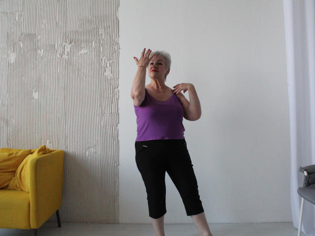 KayleSmoll, elderly woman with short white hair, purple tank top, black capri pants, standing, arms raised, yellow chair on left, white wall, light wooden floor, gray floor lamp.