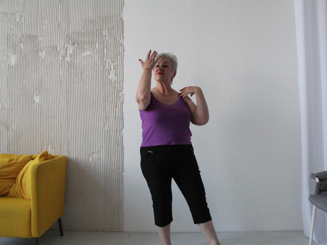 KayleSmoll, an older woman with short gray hair, purple tank top, black capris, and red nails, stands in a dance pose, arms raised, in a minimalist room.