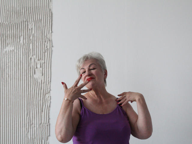 KayleSmoll, an older woman with short white hair, wearing a purple tank top, has her eyes closed and red lipstick, touching her face and shoulders with red nails.