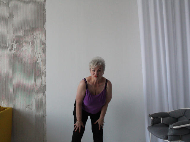 KayleSmoll has short white hair, purple tank top, black pants, and bends forward with hands on knees, showing small tits and a slightly saggy ass.