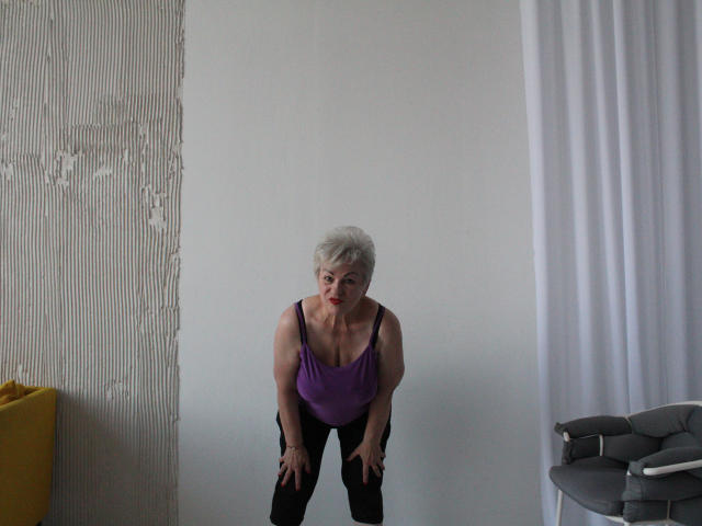 KayleSmoll, short gray hair, purple tank top, black pants, bending forward, hands on knees, showing medium-sized tits, white wall, yellow bucket, gray chair, white curtain.