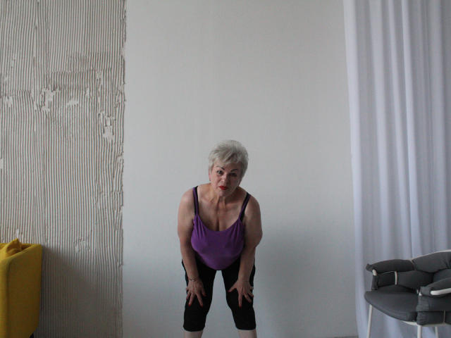 KayleSmoll, short white hair, purple tank top, black pants, bending forward, hands on knees, red lipstick, white wall, yellow chair, gray table, white curtain, looking at camera.