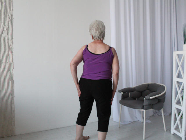 KayleSmoll has short white hair, a purple tank top, black capri pants, and stands with hands on hips, facing away, showing a slightly chubby, older ass and back.