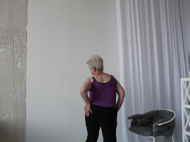 KayleSmoll, middle-aged with short gray hair, purple tank top, black pants, standing with hands on hips, facing away, showing her ass and back in a modern room.