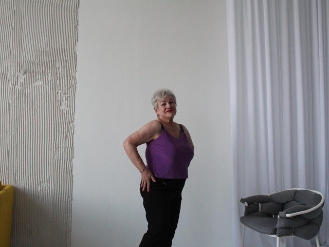 KayleSmoll, a chubby older woman with short gray hair, purple tank top, and black pants, stands with hands on hips, looking slightly to the side, in a minimalist room.