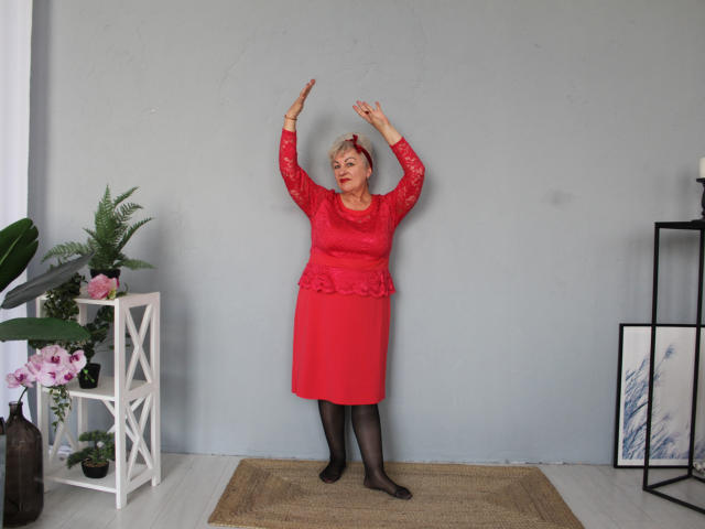 KayleSmoll stands with arms raised, wearing a red lace top and matching skirt, black tights, and red lipstick, in front of a gray wall, with a plant and shelf.
