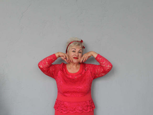 KayleSmoll with short white hair, red headband, red lace dress, and red lipstick, hands near face, standing against a plain wall, wearing a red ribbon on head.
