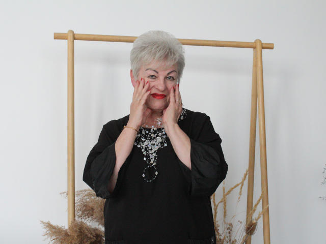 KayleSmoll has short gray hair, red lipstick, and black dress with silver sequins. Hands on face, smiling. Wooden frame behind, dried puffs to the right.