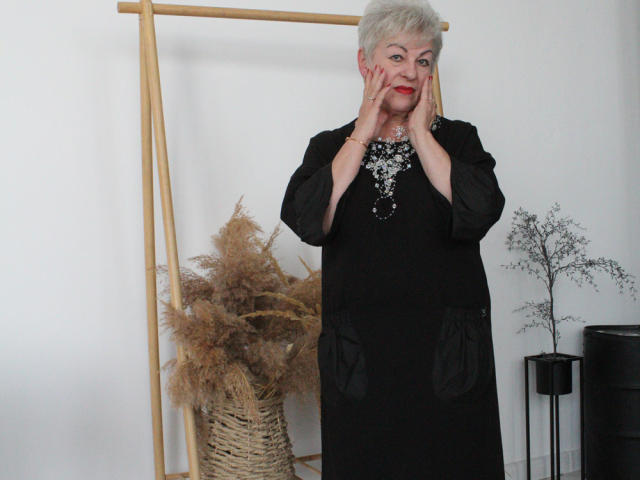 KayleSmoll, elderly woman with short white hair, red lipstick, black dress, and silver necklace, hands on face, standing in front of bamboo frame and dried pampas grass.