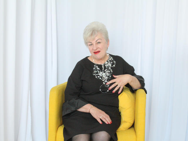 KayleSmoll with short white hair, red lipstick, black dress with sparkly chest, yellow chair, red nails, white curtain, sitting, smiling, right hand on chest, left hand on chair.