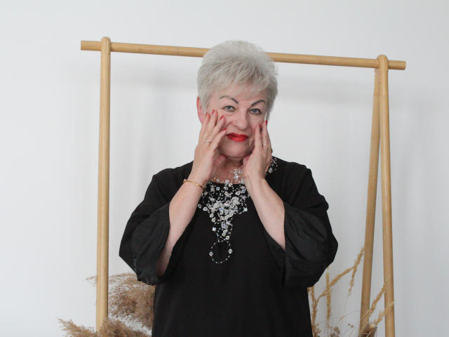 KayleSmoll with short gray hair, red lipstick, black long-sleeve shirt, hands on face, wearing a silver bead necklace, standing in front of a wooden frame, smiling.