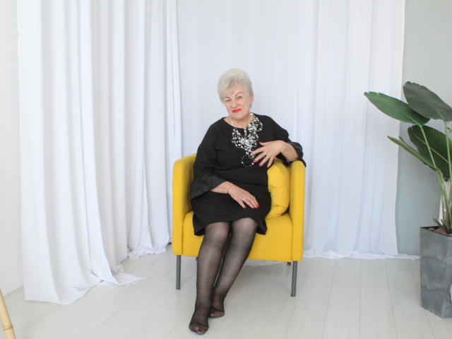 KayleSmoll, elderly woman with short white hair, red lipstick, black dress, sparkling necklace, black sheer tights, yellow chair, white curtains, sitting, right hand on chair, left hand on knee.