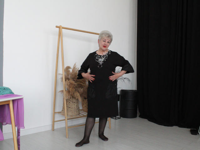 KayleSmoll stands in a black dress with tits covered by a sparkly pattern, black stockings, and hands on hips, in front of a wooden shelf with pampas grass.