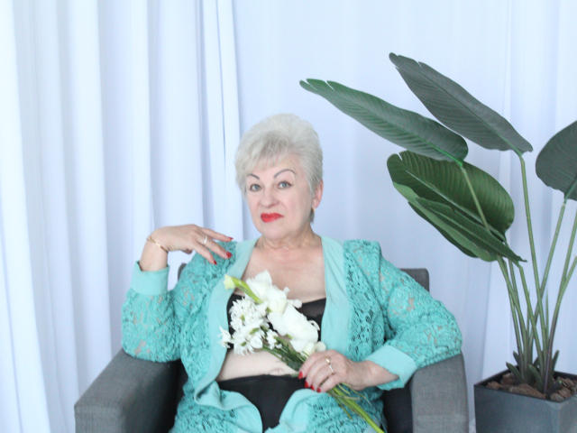 KayleSmoll, an older woman with short white hair, red lipstick, and turquoise lace outfit, sits in a gray chair, holding white flowers, with a large green plant to her right.