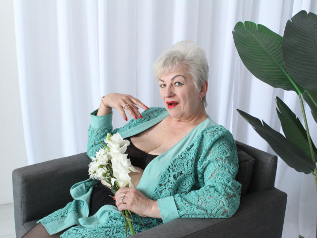 KayleSmoll with white hair, red lipstick, and teal lace robe, holding white lilies, sitting in gray chair, showing black bra and tits. Potted plant on right.
