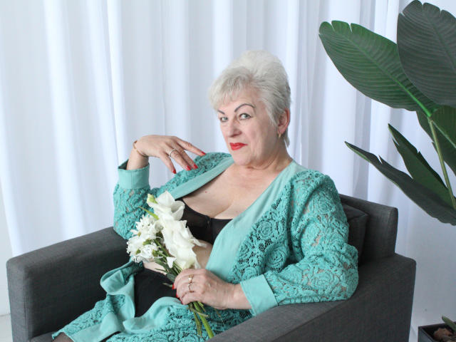 KayleSmoll with short white hair, red lipstick, green lace robe, exposing black bra and holding white flowers in gray armchair, next to large green plant.
