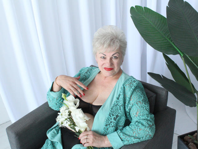 KayleSmoll, elderly woman with white hair, red lipstick, turquoise lace robe, holding white flowers, grey chair, large green plant, black bra, red nails, confident, sexy, tits visible.