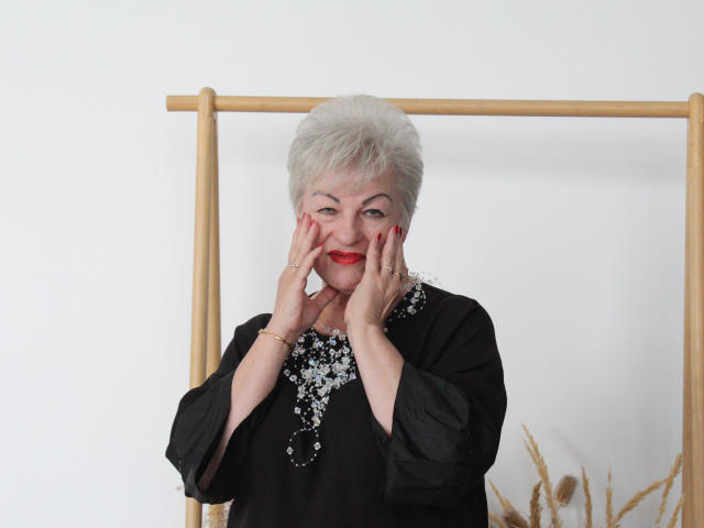 KayleSmoll with short gray hair, red lipstick, black dress, and sparkly necklace, hands on face, smiling, standing in front of a wooden frame, wearing a gold bracelet.