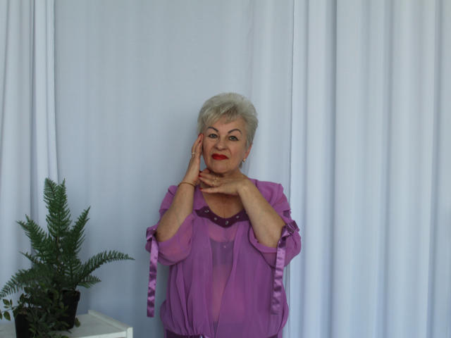 KayleSmoll with short white hair, purple sheer top, and red lipstick, hands near face, standing against white curtains, small green plant on left, looking confident and cute.