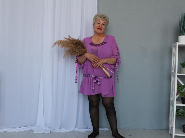 KayleSmoll stands in a purple dress holding pampas grass, with short gray hair, red lipstick, black stockings, and a gray wall behind her. She's holding a wooden stick.