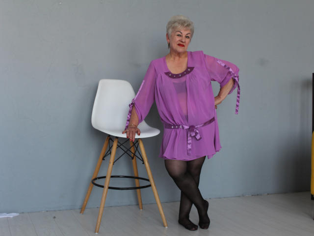 KayleSmoll with short blonde hair, purple sheer dress, black tights, standing next to white chair, hands on hips, wearing red lipstick, wooden legs on chair, gray wall.