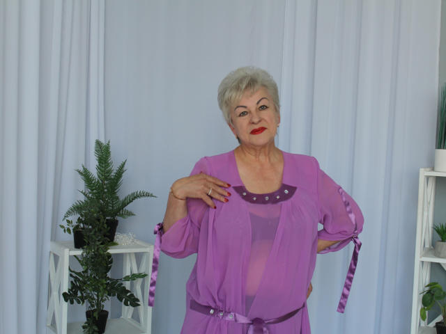 KayleSmoll with short gray hair, wearing a sheer purple robe, exposing her small, sagging tits, and a purple belt, standing in front of white curtains.