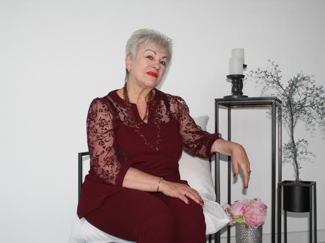 KayleSmoll with short gray hair, red lipstick, and maroon lace dress, sitting on a chair, right arm resting, left hand on pink flowers, black candle on metal stand.
