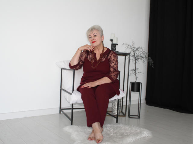 KayleSmoll, elderly woman with short gray hair, wearing red lace dress and matching pants, sitting on white chair, right hand touching chin, black curtain to the right.