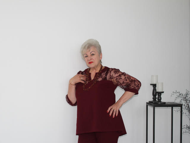 KayleSmoll has short white hair, red lipstick, and wears a maroon lace top with matching pants, standing with hand on chest, candles on a black table to the right.