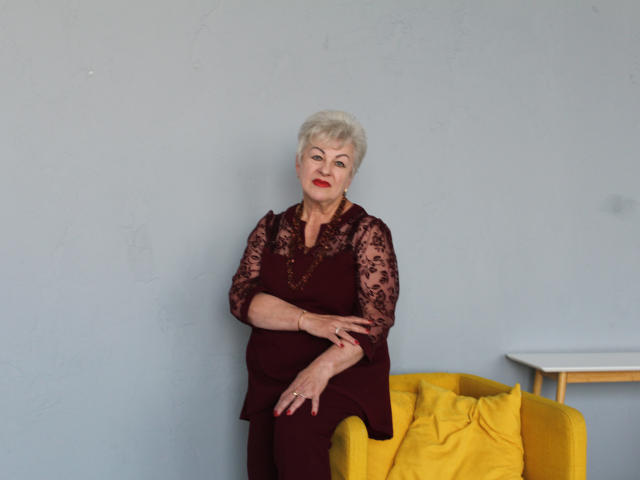 KayleSmoll, an older woman with short white hair, wearing a lace burgundy dress, stands with arms crossed, leaning on a yellow chair, looking at the camera.