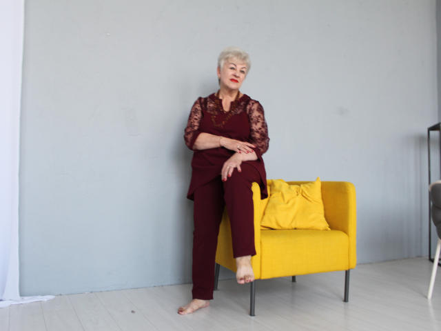 KayleSmoll, an older woman with short blonde hair, sits barefoot on a yellow chair, wearing a maroon lace top and pants, with a yellow pillow.