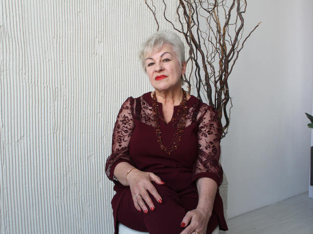 KayleSmoll, elderly woman with short white hair, red lipstick, maroon lace blouse, and gold necklace, sitting, red nail polish, hand on lap, white wall, brown branches.