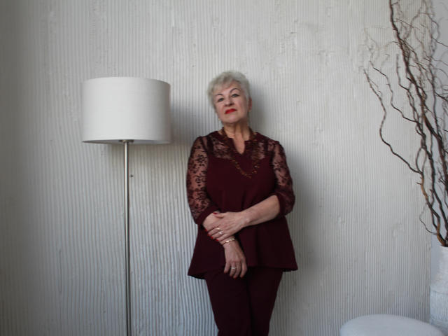 KayleSmoll, elderly woman with short white hair, red lipstick, lace sleeves, maroon top, and pants, standing, arms crossed, next to white floor lamp, and bare branch decor.