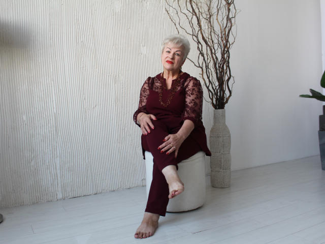 KayleSmoll, elderly woman with short white hair, wearing maroon lace dress and pants, sitting on white ottoman, right foot up, smiling, red lipstick, beige textured wall, vase with branches.