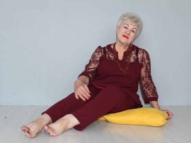KayleSmoll, an older woman with short white hair, red lipstick, and red clothes, sits on a yellow pillow, legs out, arms resting, feet bare, wearing a lace top.