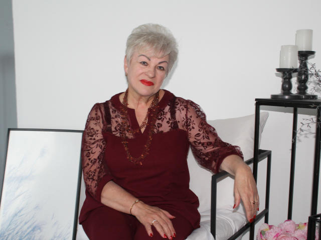 KayleSmoll, an older woman with short white hair, red lipstick, and maroon lace dress, sitting on a white chair, arms resting on black metal frame, smiling.