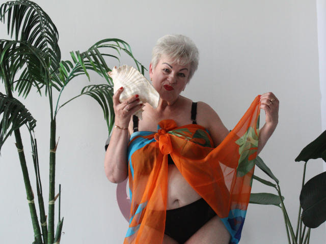 KayleSmoll, short white hair, holding shell, wearing black bra, black panties, orange and blue sheer scarf, exposing her tits, standing with green plants, smiling, red lipstick.