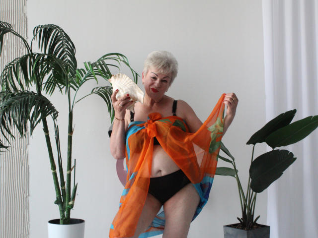 KayleSmoll with short white hair, black bra, black panties, holding orange and blue sheer scarf, showing tits, holding fan, standing by potted plants, looking at camera.
