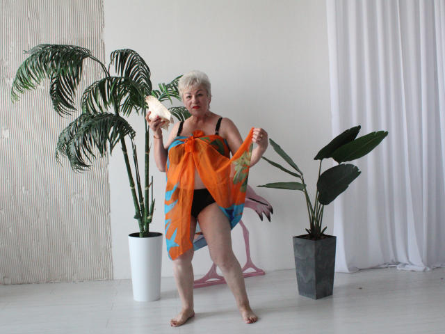 KayleSmoll, an older woman with short white hair, holding a banana, wearing a black bra and orange-blue sheer towel, standing barefoot, in front of potted plants.