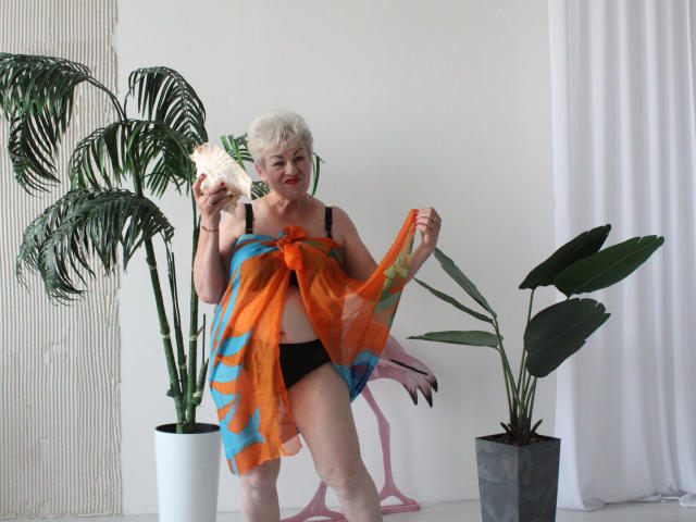 KayleSmoll with short white hair, black bra, and black panties, holding orange and blue sheer scarf, exposing her tits and pussy, standing in front of green plants.
