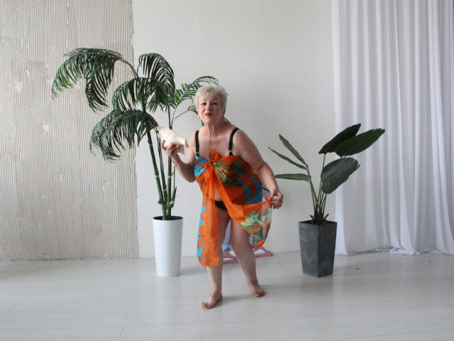 KayleSmoll with short white hair, red lipstick, and a colorful, semi-transparent orange dress, holding a white flower, standing barefoot, surrounded by potted plants in a bright room.