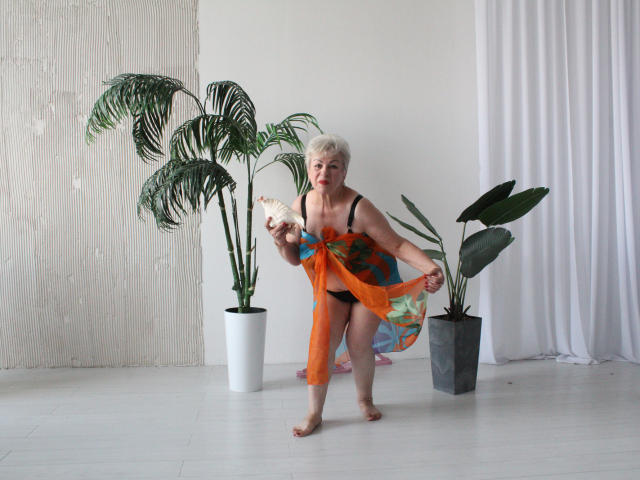 KayleSmoll with short white hair, wearing a sheer orange and blue dress, black bra, and panties, holding a white feather, standing barefoot in front of two potted plants.